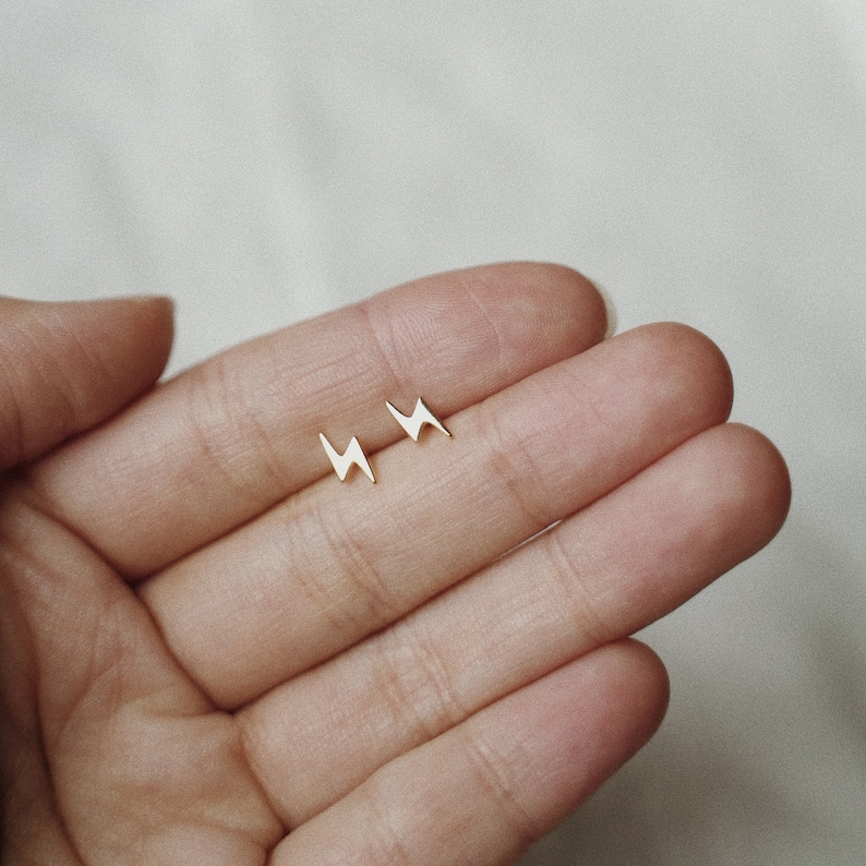 Puede incluir: Un par de peque&ntilde;os pendientes de tuerca dorados con forma de rayo. Los pendientes se muestran sobre un fondo claro, mostrando su dise&ntilde;o minimalista. Los pendientes se sostienen en una mano.