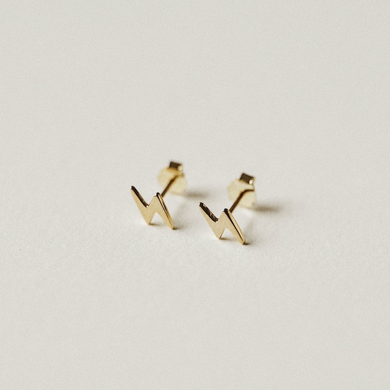 Puede incluir: Un par de pendientes dorados con forma de rayo. Los pendientes son peque&ntilde;os y minimalistas, con un dise&ntilde;o geom&eacute;trico simple. Los postes son visibles en la parte posterior de los pendientes.