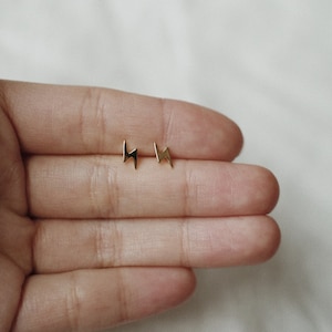 Puede incluir: Un par de peque&ntilde;os pendientes de tuerca dorados con forma de rayo. Los pendientes se muestran sobre un fondo claro. Dise&ntilde;o sencillo y minimalista.