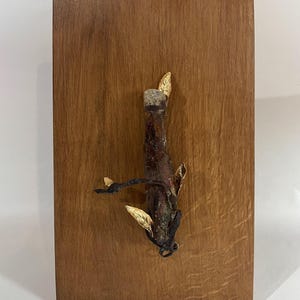 Peut inclure: Une planche de bois rectangulaire avec un grain de bois visible. Un objet organique brun foncé est fixé au bois, avec des détails dorés. L'objet ressemble à une tige de plante séchée ou à un élément naturel similaire.