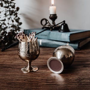 May include: Two brass chalice-shaped containers with etched floral patterns. One container stands upright and is filled with wooden matches with brown tips, while the other lies on its side, revealing a circular base with a dark brown center.