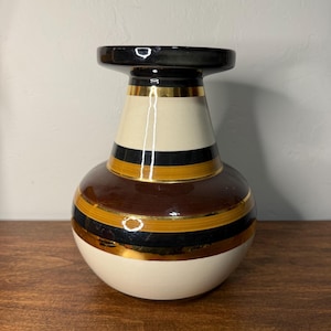 May include: A decorative ceramic vase with a black top, cream-colored midsection, and a rounded brown base. The vase features gold and black horizontal stripes. The vase is approximately 10 inches tall.