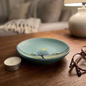 Op de afbeelding: Een kleine, ronde, lichtblauwe keramische schaal met een geschilderde landschapsscène. De scène toont een boom zonder bladeren, een gele zon en een grasveld. Een brandende waxinelichtkaars staat links van de schaal. Schildpadbrillen staan rechts.