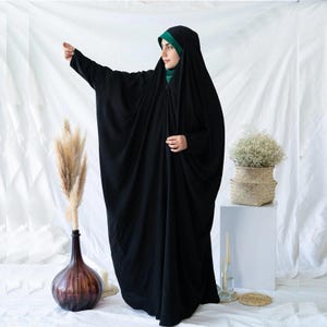 May include: A black abaya with a hood, worn over a green head covering. The abaya is a loose-fitting garment. The image is set against a white backdrop with decorative elements including a vase with dried plants and a basket of flowers.