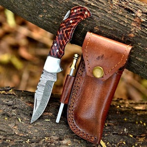 May include: A folding knife with a patterned blade and a brown and red handle, next to a brown leather sheath with a gold button closure. A small sharpening tool is also included.