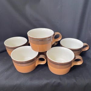 May include: A set of five ceramic mugs featuring a two-tone design with a dark brown upper band and a lighter terracotta orange lower section. Each mug has a wide, rounded body, a sturdy side handle, and a speckled off-white interior finish.