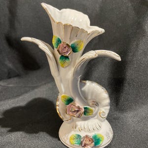 May include: A white ceramic vase with a unique, curved design. The vase features pink and gold rose accents with green and yellow leaves. The edges are trimmed with gold. The vase is approximately 7 inches tall.