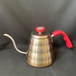 May include: A stainless steel gooseneck kettle featuring a brushed metallic finish, a curved spout, and a bright red handle. The kettle includes a matching red lid knob with a silver center, designed for precise pouring in a kitchen setting.