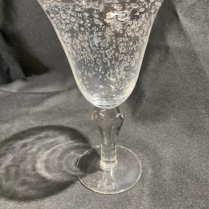 May include: A clear, stemmed wine glass with a wide bowl and a round base. The glass is decorated with small, irregular bubbles throughout the bowl. The stem has a bulbous shape, and the base is flat.