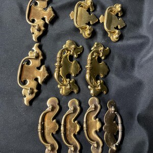 May include: A collection of nine brass drawer pulls featuring ornate, scalloped backplates and curved swinging handles. The hardware displays a polished metallic finish with intricate decorative edges and rounded mounting points for furniture attachment.