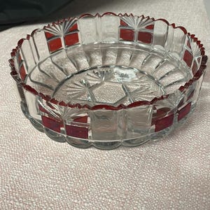 May include: A clear glass bowl with a scalloped rim and a red border. The bowl has a starburst pattern on the bottom and red rectangular accents around the rim. The bowl is likely for serving or display.
