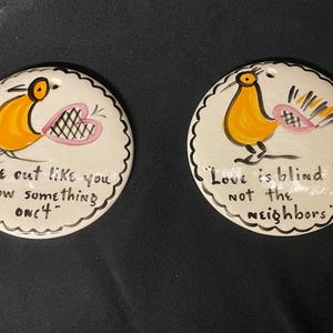 May include: Two round, white ceramic ornaments with scalloped edges. Each features a cartoon bird in yellow and black, with pink accents. One reads "Make out like you know something once." The other says "Love is blind but not the neighbors."