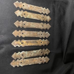 May include: Eight antique bronze-colored metal decorative plates. Each plate has a rectangular center with decorative ends and three holes for mounting. The plates show signs of age and wear, with a weathered patina.