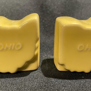 May include: Two yellow ceramic salt and pepper shakers shaped like the state of Ohio. The word "OHIO" is embossed on the front of each shaker. The shakers are on a dark gray surface.