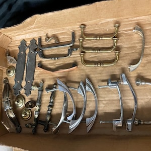 May include: A collection of vintage cabinet hardware, including handles and pulls, in various styles and finishes. The hardware is made of metal and includes brass, silver, and bronze tones. The items are arranged inside a cardboard box.