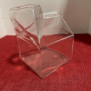 May include: A clear glass creamer shaped like a milk carton. The creamer has a rectangular base and a slanted top, with a small opening for pouring. The glass is transparent, allowing the red surface beneath to be visible.