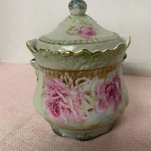 May include: A vintage, lidded ceramic jar with a light green base and lid, adorned with pink rose floral designs. The jar features gold trim and a decorative handle on the lid. The overall design is ornate and elegant.