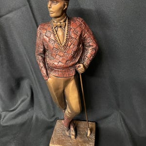 May include: Bronze sculpture of a golfer in a classic pose, wearing a cap, argyle sweater, and trousers. The figure holds a golf club, standing on a square base. The sculpture has a vintage aesthetic and is finished in shades of brown and gold.