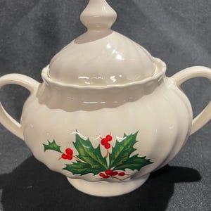 May include: Cream-colored ceramic sugar bowl with a lid and two handles. The bowl is decorated with a holly and berry design in green and red. The lid has a decorative finial.