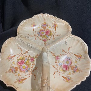 May include: A three-sectioned, off-white ceramic serving dish with a handle. The dish is decorated with floral bouquets in pink, yellow, and blue, with gold accents. The edges have a scalloped design.