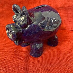 May include: A glossy, deep purple ceramic piggy bank shaped like a pig with a snout, two upright ears, and two protruding eyes. The surface has a mottled, dark blue and purple glaze with a coin slot located on the top of the back.