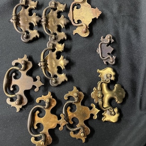 May include: A collection of antique brass drawer pulls with ornate, floral-inspired designs. The pulls feature curved handles and decorative backplates, showcasing a vintage aesthetic. The brass has a warm, aged patina, adding to their classic appeal.