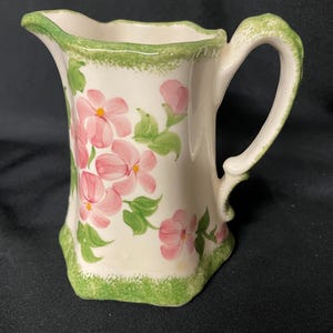 May include: A small, white ceramic pitcher with a curved handle and a spout. The pitcher is decorated with pink floral designs and green leaves. The rim and base are trimmed with green. The pitcher is approximately 6 inches tall.