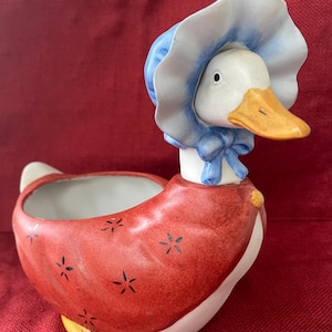 May include: A ceramic duck planter with a red body adorned with black star-shaped designs. The duck wears a blue bonnet and bow, with a white face, and an orange beak. The planter has a white interior and yellow feet.
