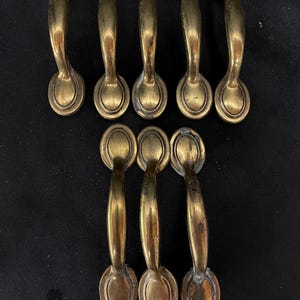 May include: A set of eight brass-toned metal drawer pulls arranged in two rows. Each handle features a curved arch design with oval-shaped mounting plates at both ends, displaying a weathered, aged finish with visible signs of patina and wear.