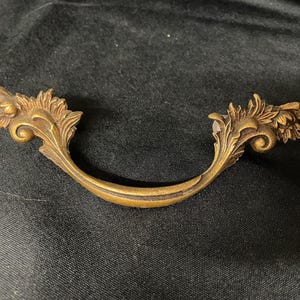 May include: A curved brass drawer pull featuring ornate, leaf-like detailing on both ends. The metal has a darkened, aged patina finish with intricate scrollwork and foliage patterns extending from the central handle grip to the mounting points.