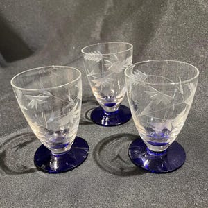 May include: Three clear glass cordial glasses with etched leaf designs. Each glass has a flared bowl and a deep blue circular base. The glasses are arranged on a dark gray surface.
