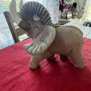 May include: A ceramic elephant figurine with large, curved tusks and a textured trunk. The elephant is primarily brown with white tusks and a small, lighter-colored elephant on its back. It is sitting on a red surface.