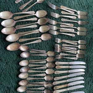 May include: An assortment of antique silverware, including spoons, forks, and knives, arranged on a green textured surface. The silverware has ornate, detailed handles and a silver finish. The collection includes various sizes of spoons, forks, and knives.