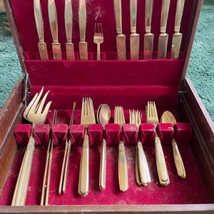 May include: A vintage silverware set in a wooden case with a red velvet interior. The set includes various forks, knives, and spoons with gold-colored handles. The case is open, displaying the neatly arranged utensils.