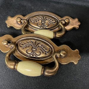 May include: Two antique bronze-toned drawer pulls with a floral design on the oval center. The handles are curved, and the center is a light yellow color. The pulls are on a dark background.