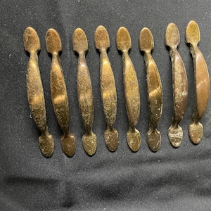 May include: A set of eight matching brass drawer pulls arranged in a row. Each handle features a curved, elongated design with a smooth center and rounded ends. The metal has a weathered, aged patina with visible texture and golden-brown tones.