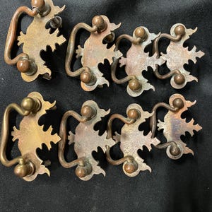 May include: A set of eight brass drawer pulls featuring a decorative backplate with a scalloped edge and a swinging bail handle. Each piece displays a tarnished metallic finish with visible signs of age and oxidation across the surface.