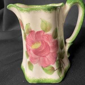 May include: A small, off-white ceramic pitcher with a pink floral design. The pitcher features a green handle and a green trim around the top and base. The floral design includes a large pink flower with a yellow center and green leaves.