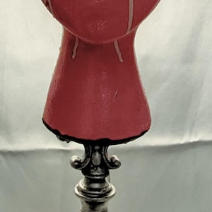 May include: A unique table lamp with a red, textured head-shaped shade featuring a white band and dripping accents. The lamp has a silver base and a dark, patterned midsection, creating a striking contrast.