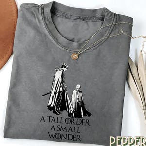 Peut inclure: T-shirt gris avec une illustration en noir et blanc de deux personnages en cape, l'un tenant une épée. Le texte "A TALL ORDER A SMALL WONDER" est imprimé en dessous. Un collier doré avec un pendentif est posé sur le t-shirt.