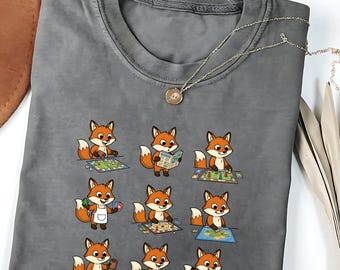 May include: A gray t-shirt featuring a playful design of cartoon foxes engaged in various board game activities. The foxes are depicted in different poses, playing games, reading rule books, and holding cards. A silver necklace with a pendant is draped across the shirt.