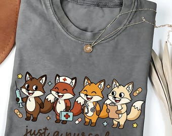 May include: Gray t-shirt featuring a cartoon design of four foxes dressed as nurses, each holding medical tools. The text "just a nurse fox" is printed below the foxes. A silver necklace with a pendant is also visible.