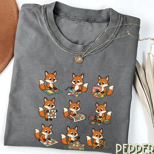 May include: Gray t-shirt featuring a repeating pattern of cartoon foxes engaged in quilting activities. Each fox is depicted with orange fur and white accents, working with sewing machines, irons, and fabric squares. A silver necklace with a pendant is draped across the shirt.