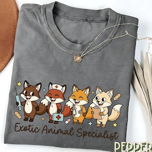 Puede incluir: Camiseta gris con una ilustración de dibujos animados de cuatro zorros en roles médicos. Los zorros sostienen una jeringa, un maletín médico, un estetoscopio y un portapapeles. El texto "Exotic Animal Specialist" está impreso debajo.