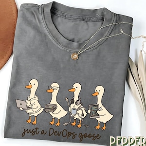 May include: Grey t-shirt with a design of four cartoon geese, each with tech accessories, and the text "just a DevOps goose." A silver necklace with a pendant is on the shirt. The t-shirt is folded.