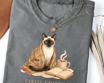 May include: Gray t-shirt featuring a Siamese cat wrapped in a blanket, with a coffee cup and an open book. The text "COFFEE, CHAPTERS and CUDDLES" is printed below the cat. A gold necklace is draped on the shirt.
