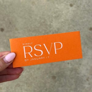 May include: An orange RSVP card with white text. The card reads "KINDLY RSVP BY JANUARY 17". The card is rectangular and held in a hand with pink nail polish.