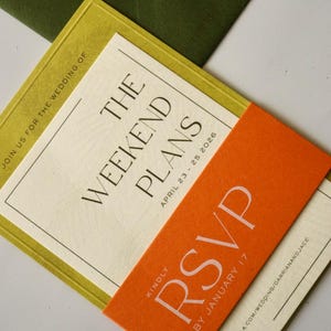 May include: A wedding invitation suite featuring a cream-colored card with black text reading THE WEEKEND PLANS APRIL 23 - 25 2026, overlaid with a bright orange RSVP band, all resting on a dark olive green envelope sealed with a gold wax stamp.