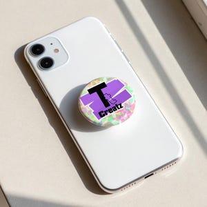 May include: A white smartphone with a circular collapsible phone grip attached to the back. The grip features a multicoloured speckled background with a purple brushstroke design and the black text T Creatz printed across the centre.