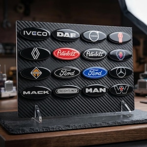 May include: A selection of oval truck badges, showcasing logos such as Iveco, DAF, Volvo, and Ford. The black badges have silver or coloured text, set against a carbon fibre-effect backdrop. The display is supported by clear acrylic stands, presented on a slate base.